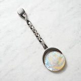 A silver-toned twisted metal spoon handle with a round iridescent mother-of-pearl shell bowl, displayed diagonally on a light gray marble surface.