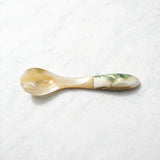 A small mother-of-pearl spoon with a warm gold-beige handle and a green-blue iridescent end, resting on a light gray marble surface.