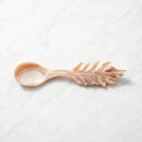 A decorative spoon with a leaf-shaped handle, crafted from a warm peachy-pink resin with a glossy finish, resting on a light gray marble surface.