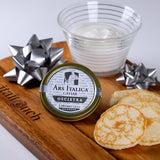 A tin of Ars Italica Oscietra Classic caviar is placed on a wooden platter alongside a glass of cream, two mother-of-pearl spoons, and a stack of pancakes.